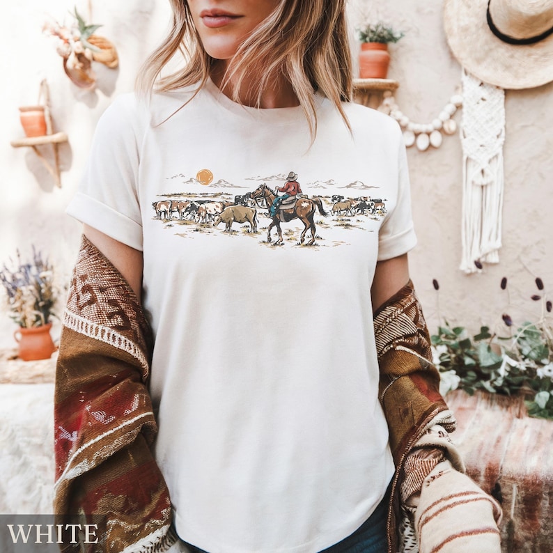 May include: White t-shirt featuring a Western-themed graphic. The design depicts a cowboy on horseback herding cattle under a setting sun. The word "WHITE" is printed at the bottom of the shirt.
