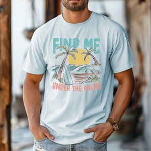 May include: A light blue t-shirt with a graphic print of palm trees, waves, and a surfboard. The text "Find Me Under The Palms" is printed on the shirt.