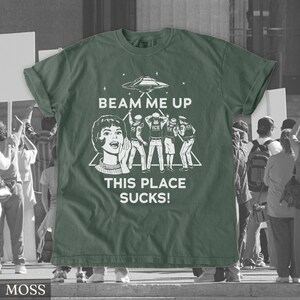 May include: A moss green t-shirt with a white graphic. The graphic includes a retro illustration of a woman, a UFO, and figures, with the text "BEAM ME UP THIS PLACE SUCKS!". The shirt has rolled-up sleeves.