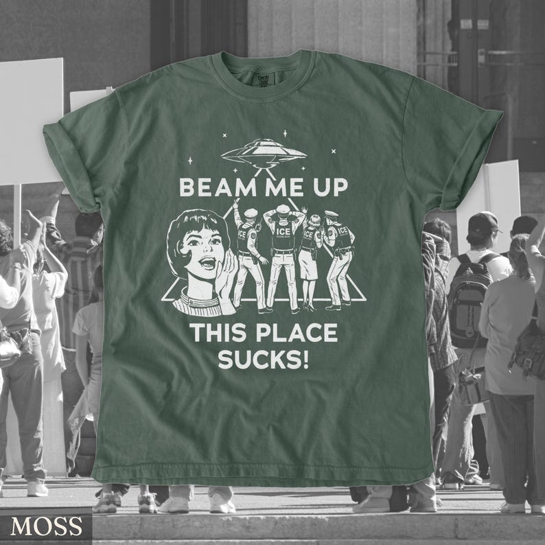 May include: A moss green t-shirt with a white graphic. The graphic includes a retro illustration of a woman, a UFO, and figures, with the text "BEAM ME UP THIS PLACE SUCKS!". The shirt has rolled-up sleeves.