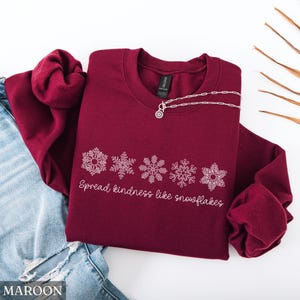 May include: A maroon sweatshirt with white snowflake designs and the text "Spread kindness like snowflakes." A silver necklace with a pendant is draped on the sweatshirt. The sweatshirt is folded on top of blue jeans.