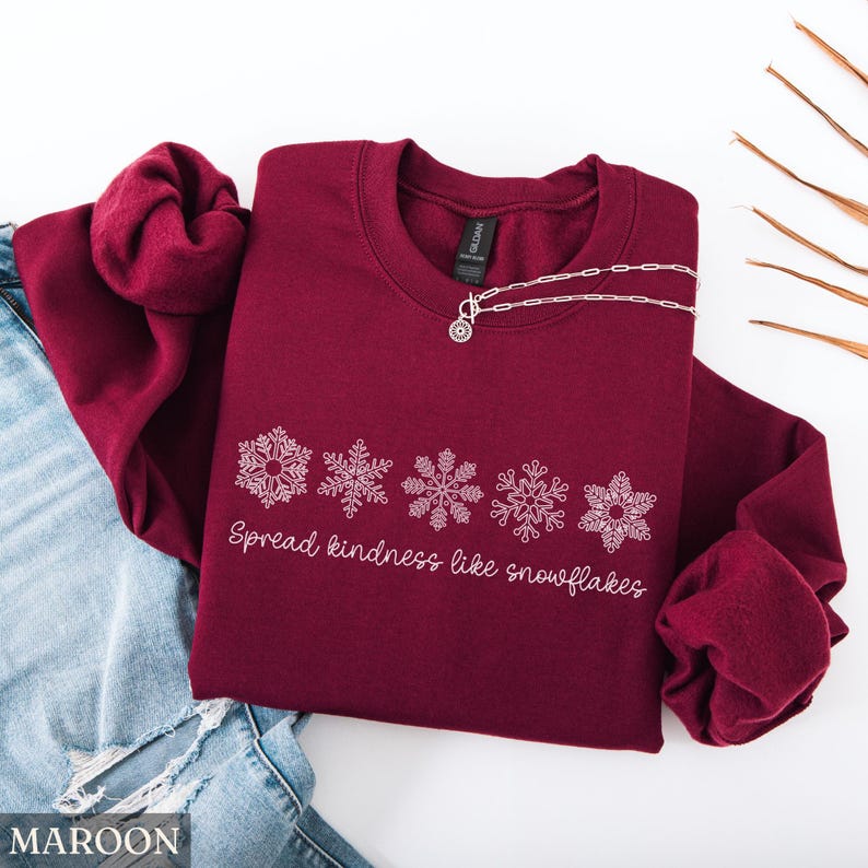 May include: A maroon sweatshirt with white snowflake designs and the text "Spread kindness like snowflakes." A silver necklace with a pendant is draped on the sweatshirt. The sweatshirt is folded on top of blue jeans.