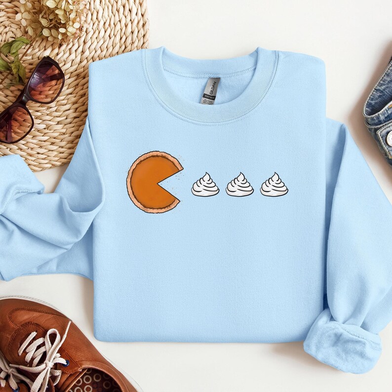 Pode incluir: Uma camisa de frio azul claro com um gr&aacute;fico de uma fatia de torta de ab&oacute;bora com uma mordida tirada, perseguindo tr&ecirc;s colheres de sopa de chantilly.
