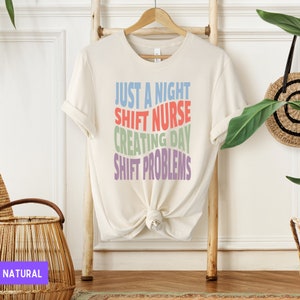 May include: A white t-shirt with the text "Just a night shift nurse creating day shift problems" in rainbow colours.