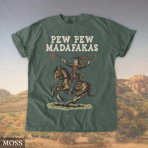 Puede incluir: Camiseta verde musgo con el texto "PEW PEW MADAFAKAS" sobre un vaquero de dibujos animados montado a caballo y disparando pistolas. El vaquero lleva una bufanda y un sombrero rojos. El caballo es marr&oacute;n.