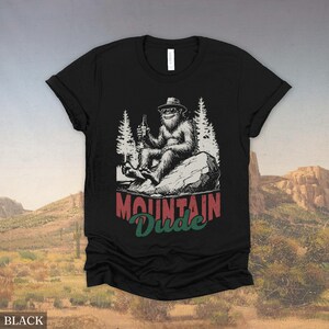 May include: Black t-shirt featuring a Bigfoot graphic. The illustration shows Bigfoot wearing a hat and sunglasses, holding a bottle, and sitting on a rock. The text "Mountain Dude" is below the graphic in red and green.