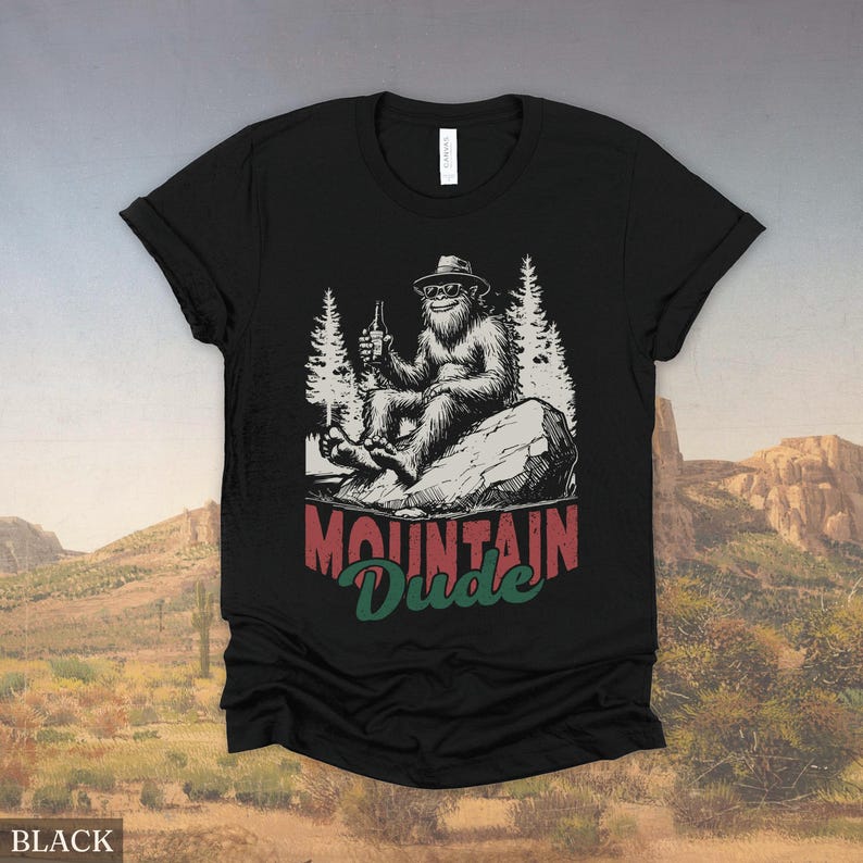 May include: Black t-shirt featuring a Bigfoot graphic. The illustration shows Bigfoot wearing a hat and sunglasses, holding a bottle, and sitting on a rock. The text "Mountain Dude" is below the graphic in red and green.