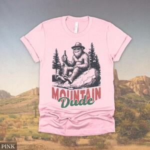 May include: Pink t-shirt featuring a Bigfoot graphic holding a beer bottle, seated on a rock with pine trees in the background. The text "MOUNTAIN Dude" is printed below the graphic in red and green.