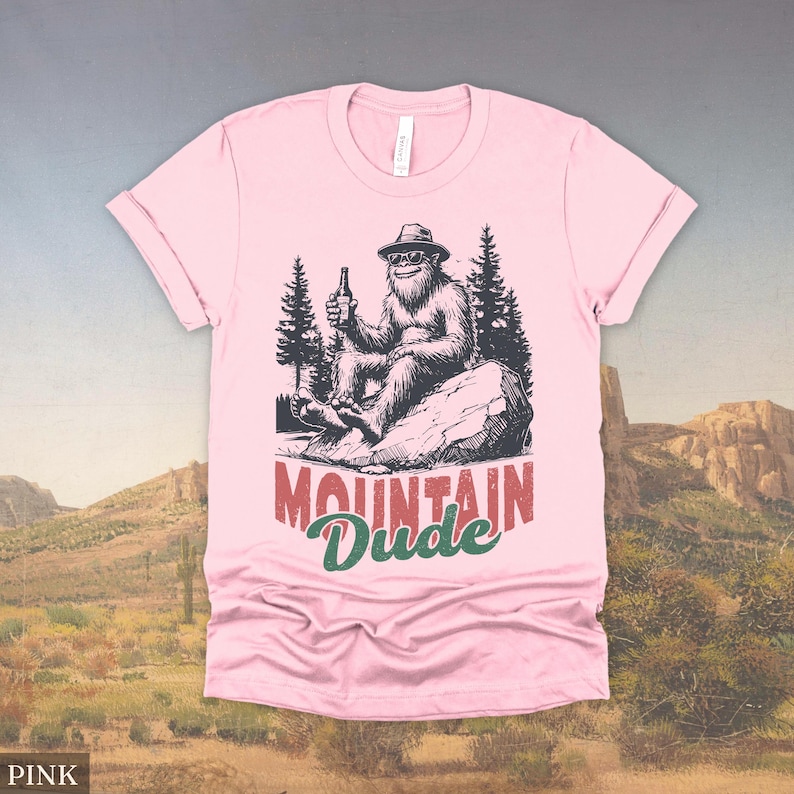 May include: Pink t-shirt featuring a Bigfoot graphic holding a beer bottle, seated on a rock with pine trees in the background. The text "MOUNTAIN Dude" is printed below the graphic in red and green.