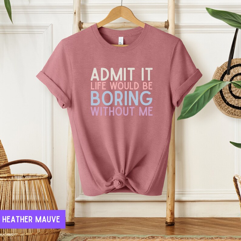 May include: Heather mauve t-shirt with the text "ADMIT IT LIFE WOULD BE BORING WITHOUT ME" in white, light blue, and purple. The shirt is knotted at the waist and hangs on a wooden hanger. The background includes a wooden ladder and decorative items.