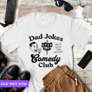 May include: White t-shirt with a black graphic design of a retro comedy club sign that says "Dad Jokes Comedy Club" with a cartoon man holding a microphone and a sign that says "Open 24/7". The text below the sign says "When the jokes are so bad they're good."