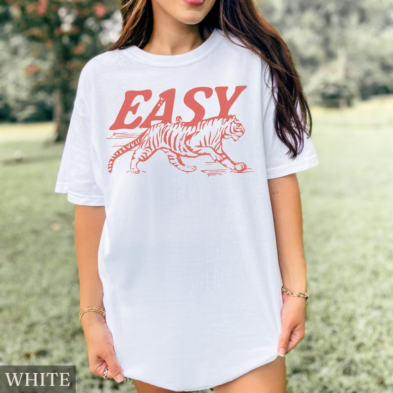 Op de afbeelding: Wit T-shirt met het woord "EASY" in rode letters boven een rode tijgerafbeelding. De tijger wordt lopend afgebeeld, met zichtbare strepen. Het shirt heeft een casual stijl.