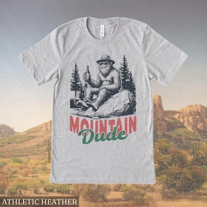May include: Athletic heather gray t-shirt with a graphic of a Bigfoot-like creature wearing sunglasses and a hat, holding a bottle. The text "Mountain Dude" is printed below the graphic.