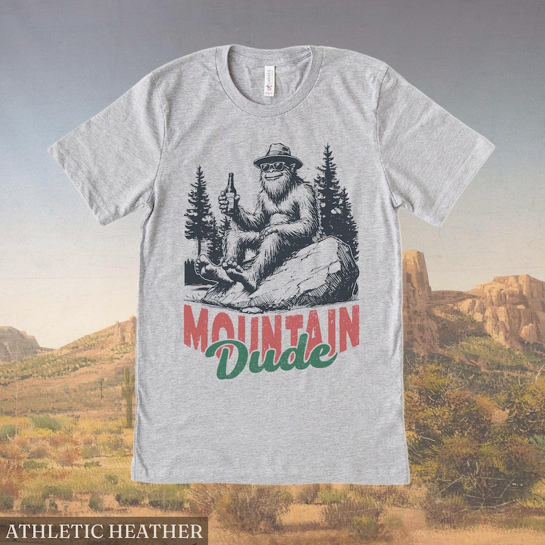 May include: Athletic heather gray t-shirt with a graphic of a Bigfoot-like creature wearing sunglasses and a hat, holding a bottle. The text "Mountain Dude" is printed below the graphic.