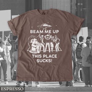 May include: Espresso-coloured t-shirt with a graphic featuring a retro-style woman, a UFO, and figures with the text "BEAM ME UP THIS PLACE SUCKS!". The shirt is a solid colour with a crew neck.