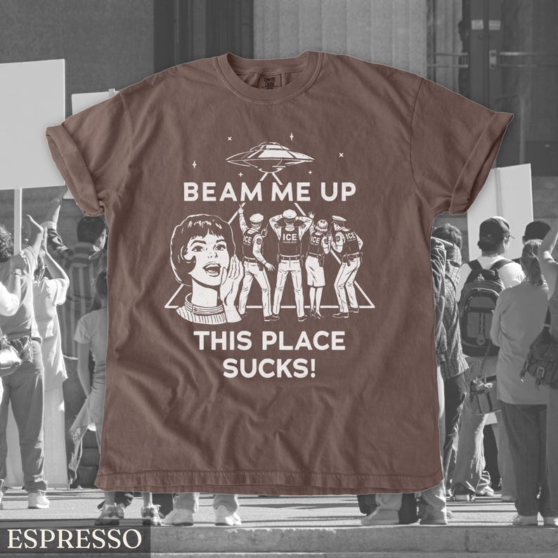 May include: Espresso-coloured t-shirt with a graphic featuring a retro-style woman, a UFO, and figures with the text "BEAM ME UP THIS PLACE SUCKS!". The shirt is a solid colour with a crew neck.