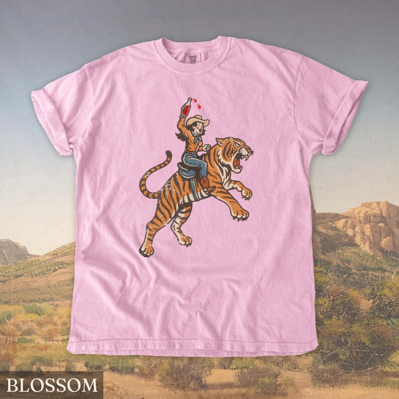 May include: Pink t-shirt featuring a graphic of a person riding a tiger, holding a bottle. The person is wearing a cowboy hat and jeans. The tiger is orange and black. The word "BLOSSOM" is printed at the bottom.