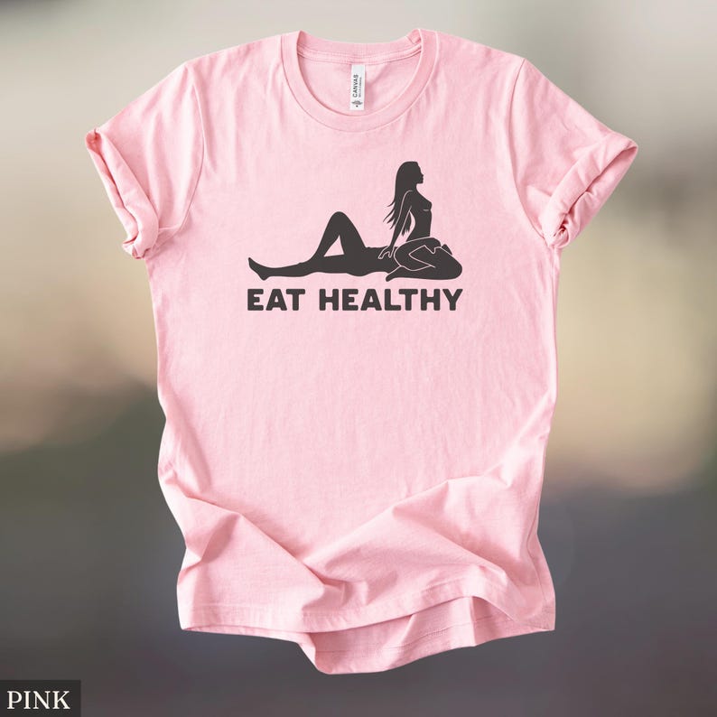 May include: Pink t-shirt with rolled-up sleeves. The shirt features a black graphic of a silhouette of a person reclining with another person sitting on top. The text "EAT HEALTHY" is printed below the graphic.