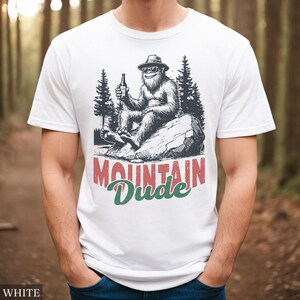 May include: White t-shirt with a graphic of a Bigfoot-like creature wearing sunglasses and a hat, holding a bottle. The text "MOUNTAIN Dude" is printed below the graphic in red and green. The shirt is made of a soft material.