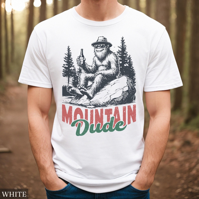 May include: White t-shirt with a graphic of a Bigfoot-like creature wearing sunglasses and a hat, holding a bottle. The text "MOUNTAIN Dude" is printed below the graphic in red and green. The shirt is made of a soft material.
