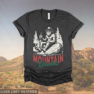 May include: Dark grey heather t-shirt featuring a graphic of a Bigfoot character wearing sunglasses and a hat, holding a beer bottle, and sitting on a rock. The text "MOUNTAIN Dude" is below the graphic.