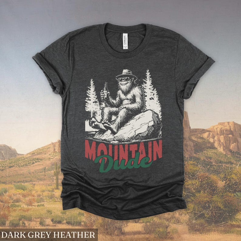 May include: Dark grey heather t-shirt featuring a graphic of a Bigfoot character wearing sunglasses and a hat, holding a beer bottle, and sitting on a rock. The text "MOUNTAIN Dude" is below the graphic.