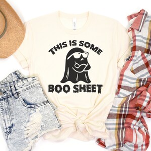 May include: A cream-colored t-shirt with a black ghost wearing sunglasses and the text "This is some boo sheet".