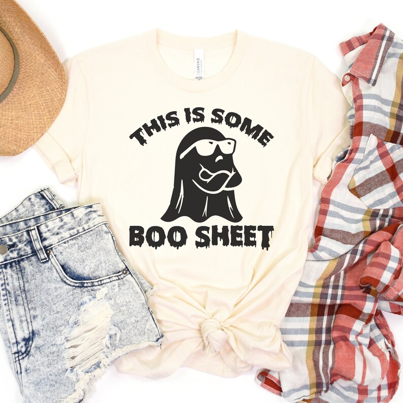 May include: A cream-colored t-shirt with a black ghost wearing sunglasses and the text "This is some boo sheet".