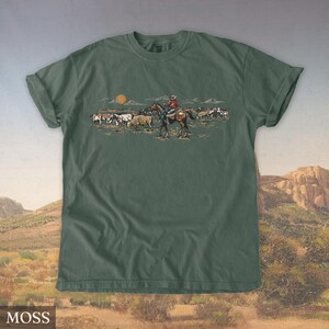 May include: Moss green t-shirt featuring a Western-themed illustration. The graphic depicts a cowboy on horseback herding cattle under a setting sun. The word "MOSS" is printed at the bottom of the shirt.