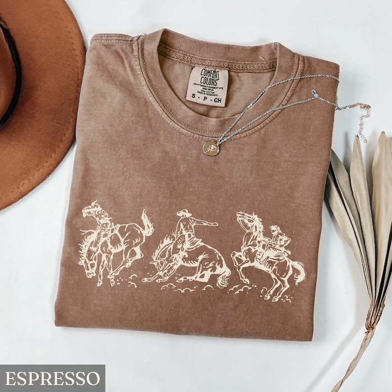Puede incluir: Camiseta color espresso con cuello redondo y un gr&aacute;fico de vaqueros montando caballos salvajes. La camiseta tiene un aspecto vintage. Un collar plateado con un peque&ntilde;o colgante est&aacute; sobre la camiseta.
