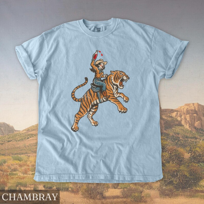 May include: Light blue chambray t-shirt featuring a graphic of a person riding a tiger, holding a bottle. The tiger is orange and black, and the person is wearing a cowboy hat. The word "CHAMBRAY" is printed at the bottom.
