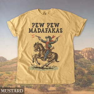 Puede incluir: Camiseta de color mostaza con un dibujo de un vaquero disparando dos pistolas a caballo. El texto "PEW PEW MADAF*KAS" est&aacute; impreso encima de la ilustraci&oacute;n. La camiseta tiene cuello redondo y mangas cortas.