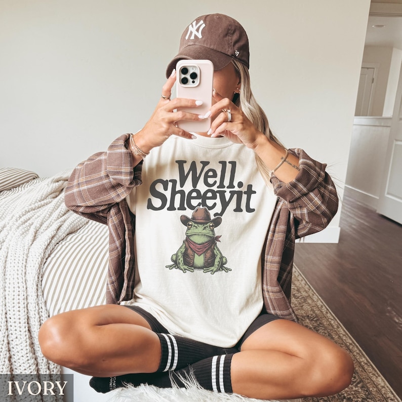 May include: Ivory-colored t-shirt with the text "Well. Sheeeyit" above a cartoon frog wearing a cowboy hat and vest. The frog has a red bandana. The person is wearing a brown cap and a brown plaid shirt.