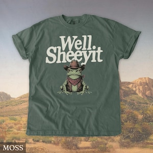 May include: Moss green t-shirt with the text "Well. Sheeeyit" in white. The shirt features a cartoon frog wearing a cowboy hat, vest, and red bandana. The background shows a desert landscape.
