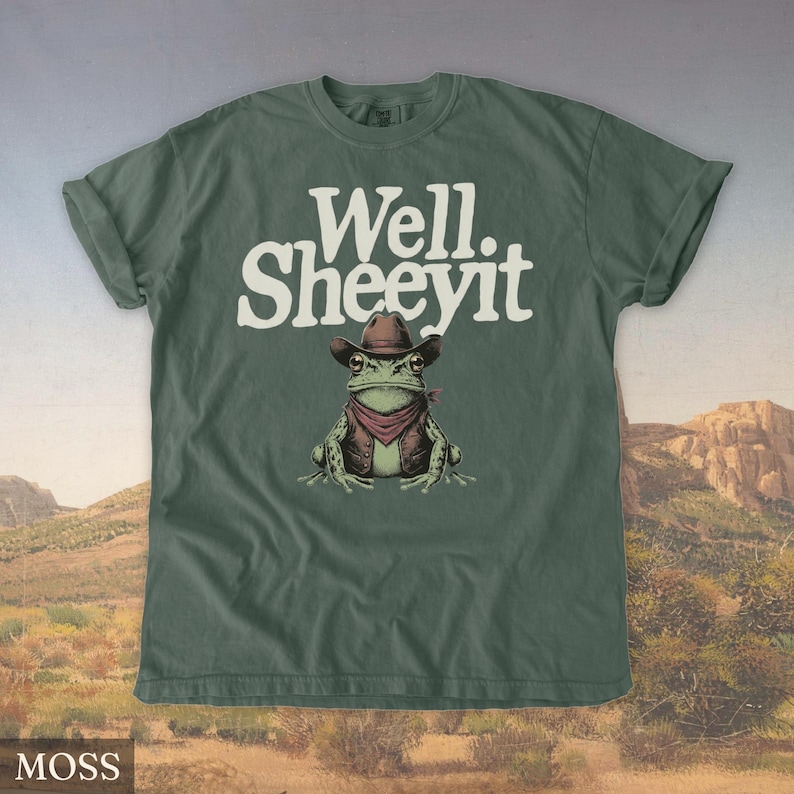 May include: Moss green t-shirt with the text "Well. Sheeeyit" in white. The shirt features a cartoon frog wearing a cowboy hat, vest, and red bandana. The background shows a desert landscape.
