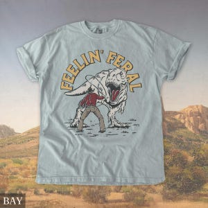 May include: Light blue t-shirt with a graphic of a cowboy roping a Tyrannosaurus Rex. The text "FEELIN' FERAL" is arched above the dinosaur. The shirt has rolled-up sleeves and a crew neck.
