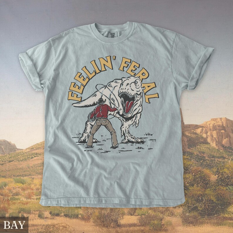 May include: Light blue t-shirt with a graphic of a cowboy roping a Tyrannosaurus Rex. The text "FEELIN' FERAL" is arched above the dinosaur. The shirt has rolled-up sleeves and a crew neck.