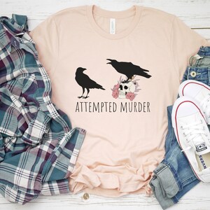 以下が含まれることがあります： 2 羽の黒いカラスが描かれたライトピンクのTシャツ。1 羽はピンクの花で飾られた頭蓋骨の上に止まっています。「ATTEMPTED MURDER」の文字が下にプリントされています。シャツはチェック柄のシャツとジーンズと組み合わせています。