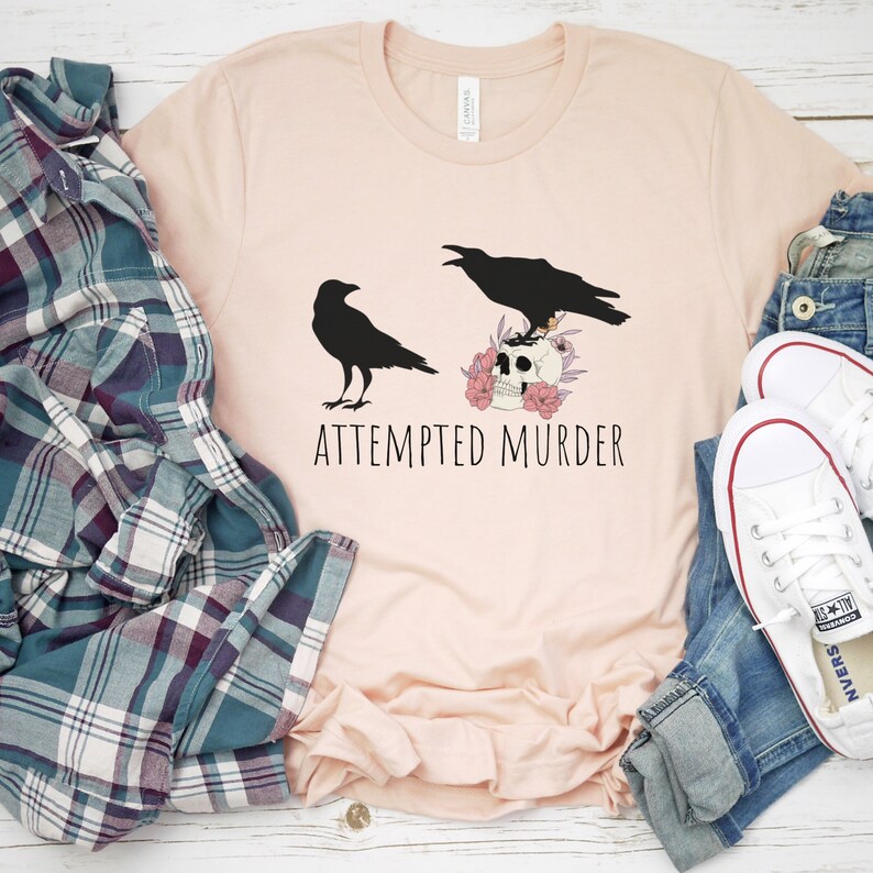 以下が含まれることがあります： 2 羽の黒いカラスが描かれたライトピンクのTシャツ。1 羽はピンクの花で飾られた頭蓋骨の上に止まっています。「ATTEMPTED MURDER」の文字が下にプリントされています。シャツはチェック柄のシャツとジーンズと組み合わせています。