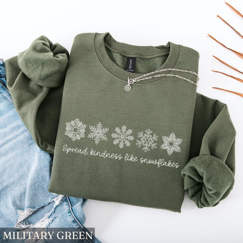 May include: Military green sweatshirt with white snowflake design and the text "Spread kindness like snowflakes." A silver necklace rests on the sweatshirt. The sweatshirt is folded on top of blue jeans.