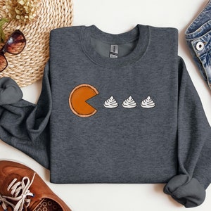 Pode incluir: Uma camisa de moletom cinza escura com um gr&aacute;fico de uma fatia de torta de ab&oacute;bora comendo tr&ecirc;s mini sobremesas de chantilly. O gr&aacute;fico est&aacute; no estilo do videogame Pac-Man.