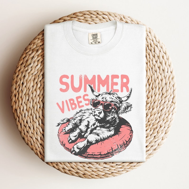 May include: White t-shirt with a graphic of a Highland cow wearing sunglasses and relaxing on a pink inflatable ring. The text "SUMMER VIBES" is printed above the cow.