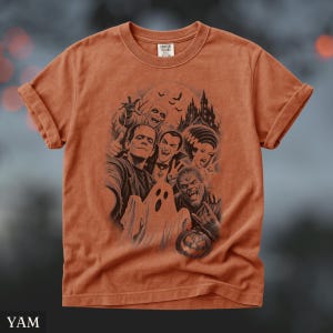 May include: A burnt orange short-sleeve t-shirt featuring a dark graphic print of classic horror movie monsters including Frankenstein's monster, a vampire, a werewolf, a ghost, and a mummy in front of a haunted castle with bats and a pumpkin.