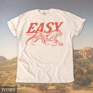 Op de afbeelding: Ivoor t-shirt met het woord "EASY" in rode blokletters boven een rode tijgerillustratie. De tijger wordt afgebeeld in een loopstand met gedetailleerde strepen. Het shirt is gemaakt van een zacht materiaal.