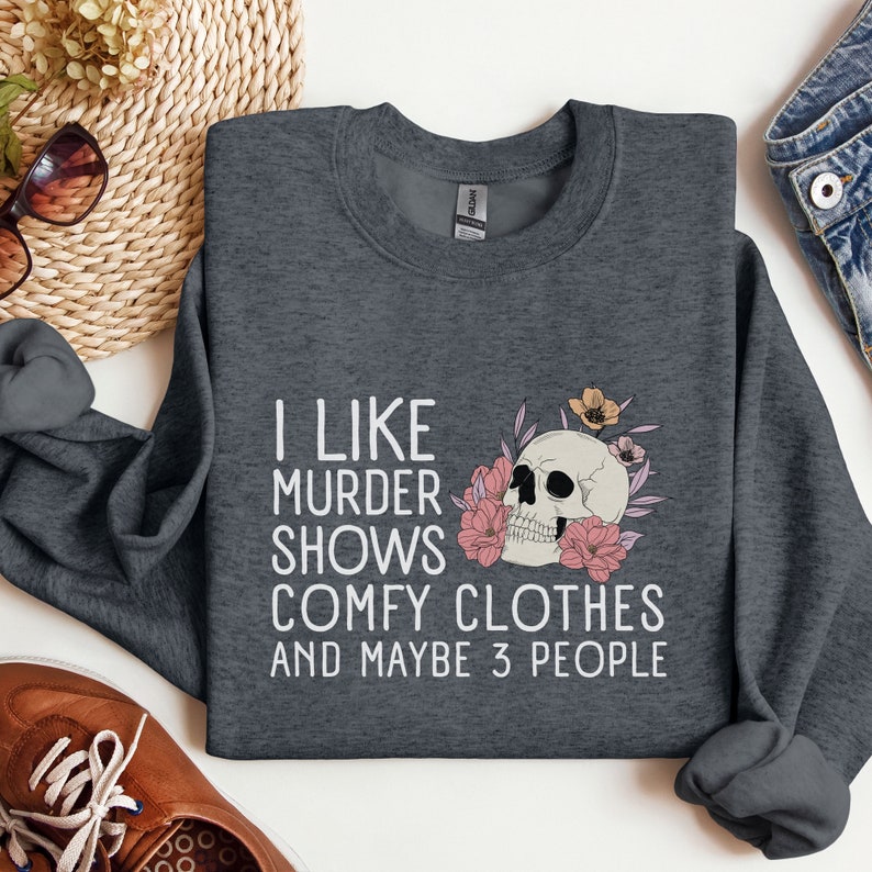 Op de afbeelding: Donkergrijze trui met een witte afbeelding die zegt "Ik hou van moordshows, comfortabele kleding en misschien 3 mensen". De afbeelding bevat ook een schedel met bloemen.