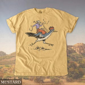 May include: Mustard-colored t-shirt featuring a cartoon illustration of a person riding a roadrunner. The rider wears a cowboy hat and holds a lasso. The roadrunner is blue, white, and black, with a red crest. The word "MUSTARD" is printed at the bottom.