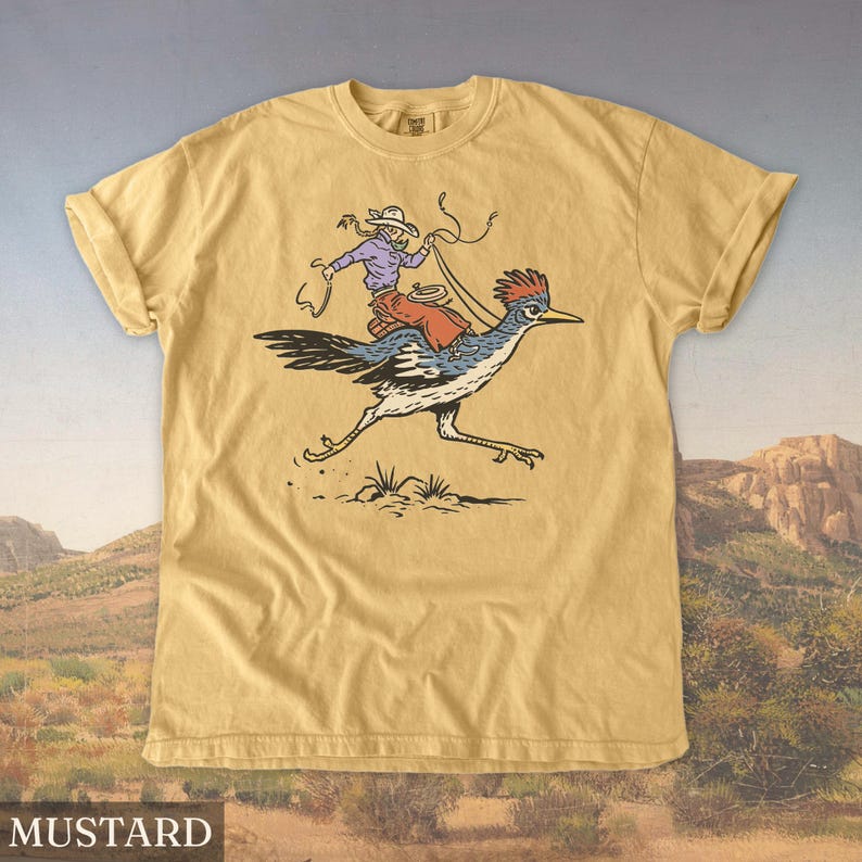 May include: Mustard-colored t-shirt featuring a cartoon illustration of a person riding a roadrunner. The rider wears a cowboy hat and holds a lasso. The roadrunner is blue, white, and black, with a red crest. The word "MUSTARD" is printed at the bottom.