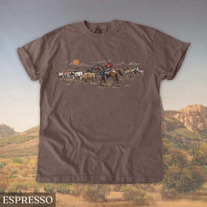 May include: Espresso-colored t-shirt featuring a Western-themed graphic. The design depicts a cowboy on horseback herding cattle under a setting sun. The shirt's color is a rich, dark brown, and the graphic is detailed with various colors.