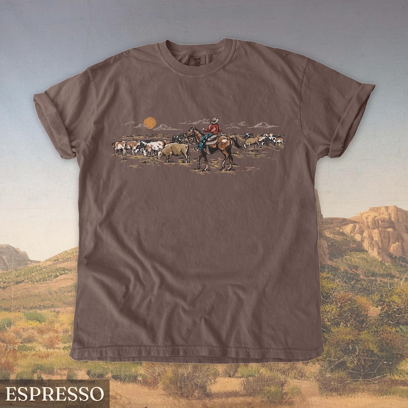 May include: Espresso-colored t-shirt featuring a Western-themed graphic. The design depicts a cowboy on horseback herding cattle under a setting sun. The shirt's color is a rich, dark brown, and the graphic is detailed with various colors.