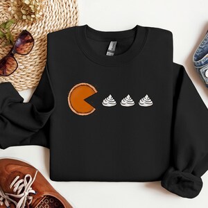 Pode incluir: Camisola de moletom preta com um gr&aacute;fico de uma fatia de torta de ab&oacute;bora comendo tr&ecirc;s marshmallows brancos. O gr&aacute;fico est&aacute; no estilo do videogame Pac-Man.